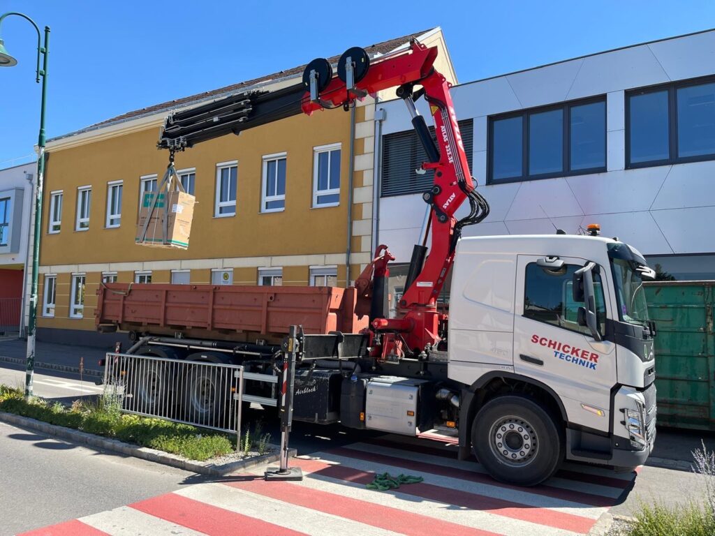 Lkw mit Kran hebt Karton vor Gebäude.